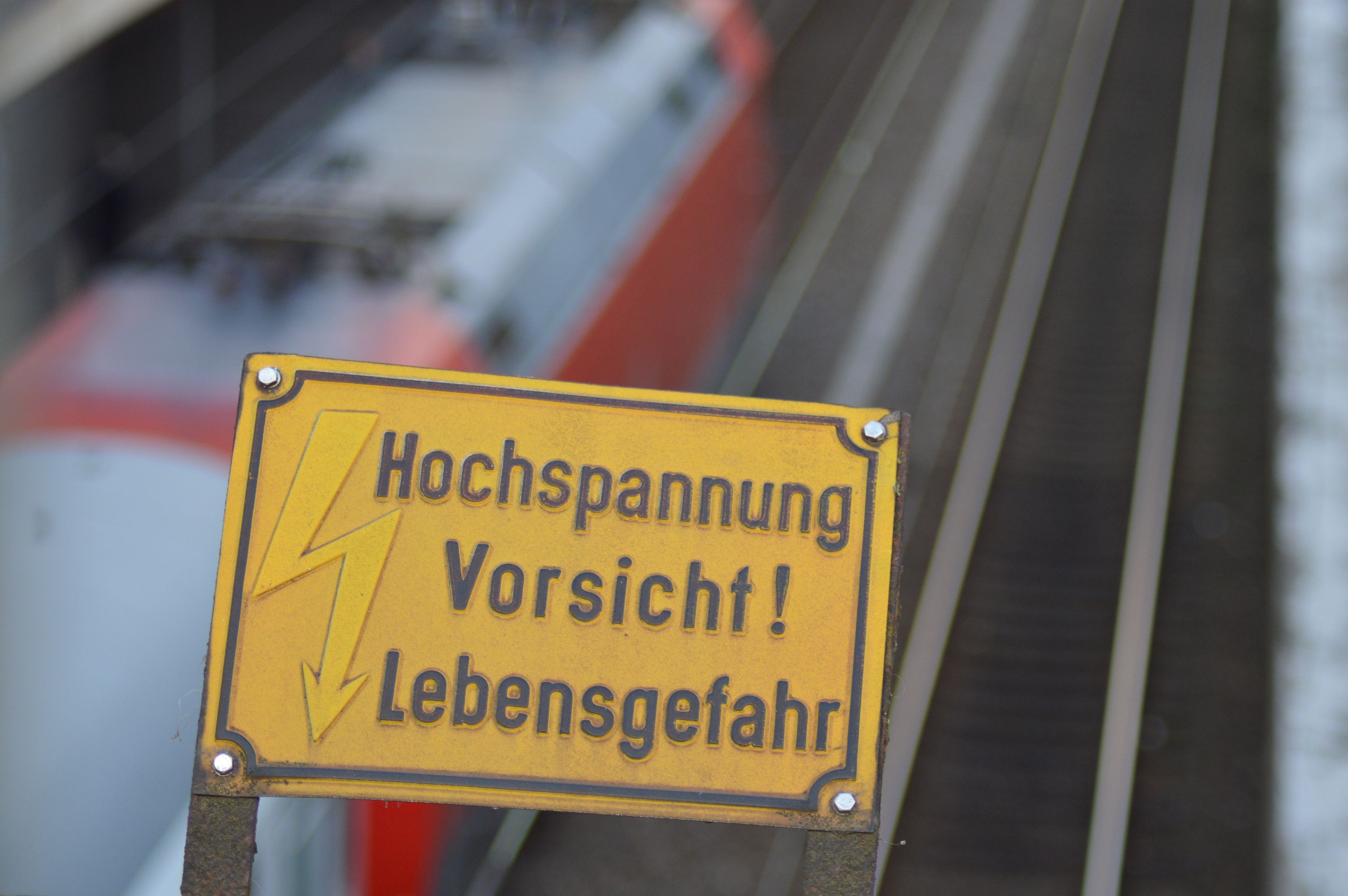 Symbolbild: Warnhinweis über der Oberleitung in einem Bahnhof. In hintergrund fährt ein Zug vorbei. (Foto: © Bahnblogstelle)
